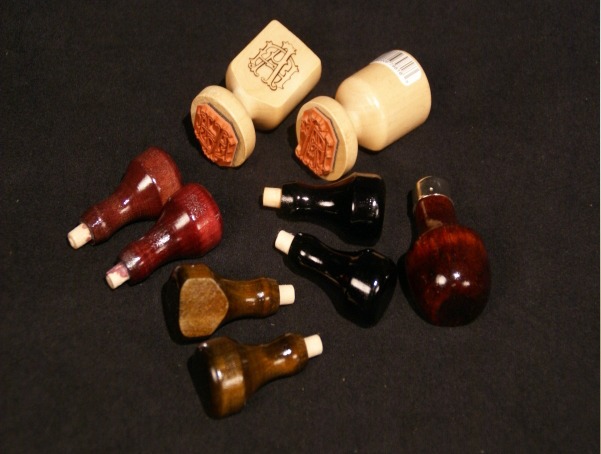 Nine custom wooden stamp handles in various finishes, including clear, mahogany stain, black paint, and walnut stain, designed for bulk manufacturing.